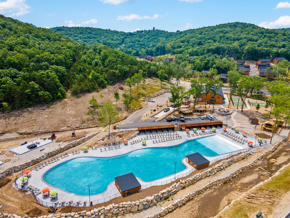 A bright, cheerful shot of a large swimming pool surrounded by lush greenery, a poolside lounge area, a wooden deck, and colorful umbrellas. The setting feels inviting, with space for families to relax, swim, and gather together outdoors.
