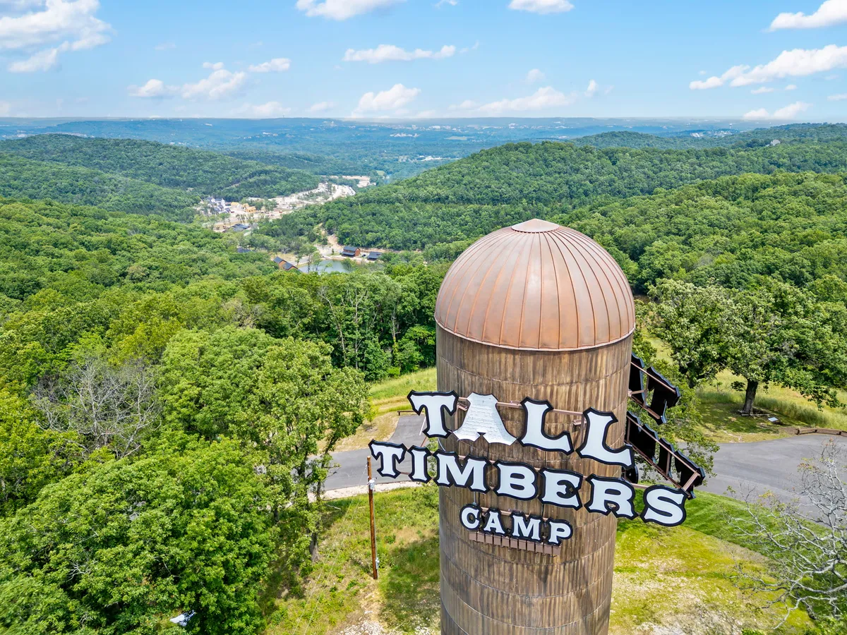 A wide view of the Tall Timbers Camp sign surrounded by scenic mountains, lush trees, and a glimpse of water in the distance. The landscape is vibrant and welcoming, instantly establishing the camp's proximity to nature and adventure.