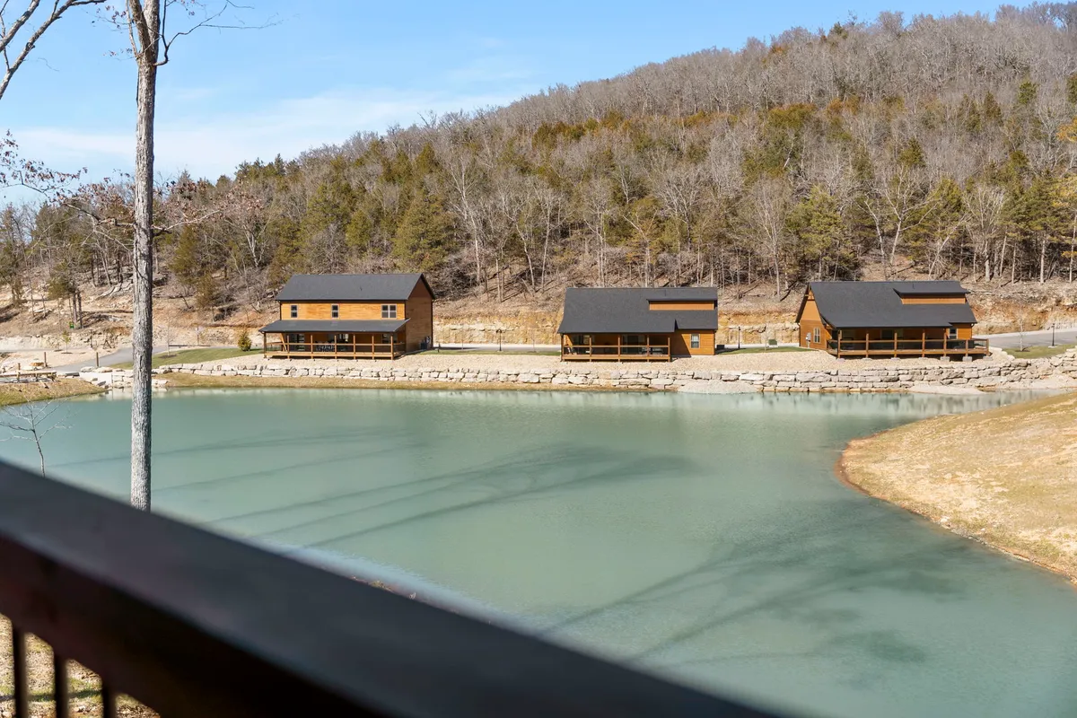 This scenic view shows three wooden cabins nestled beside a tranquil lake, surrounded by lush trees. The calm water reflects the sunrise, evoking a sense of peace and privacy, while also hinting at fishing and lakeside relaxation opportunities.