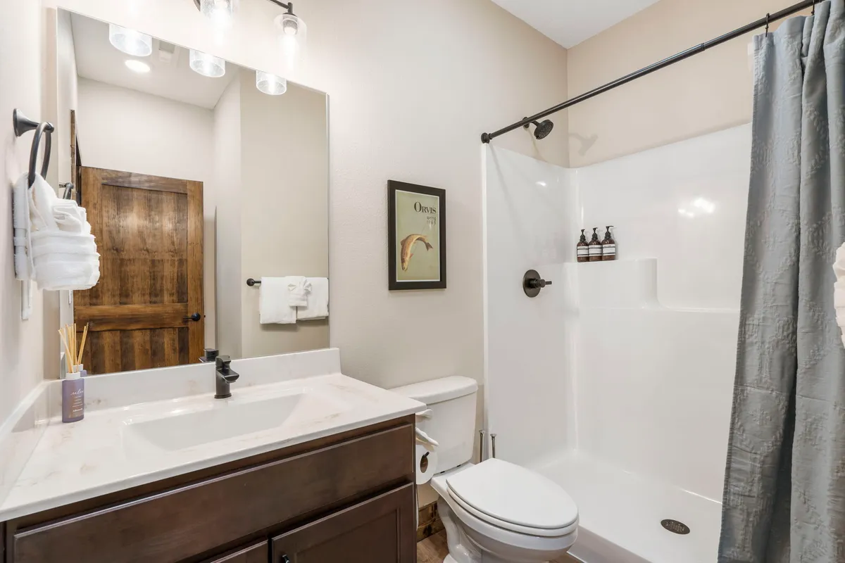 A modern bathroom with a vanity sink, spacious shower area, wooden door, and decorative framed artwork. The bathroom is bright, clean, and inviting, signaling comfort and convenience for guests.