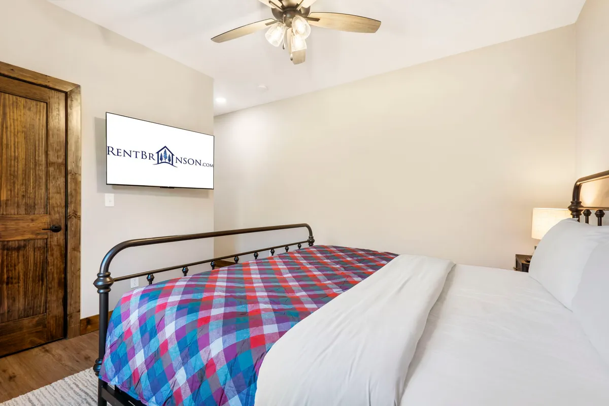 A cozy bedroom featuring a bed with colorful plaid bedding, a TV mounted on the wall, a wooden door, and a ceiling fan. The room is softly illuminated by natural light, creating a welcoming atmosphere ideal for relaxation and leisure.