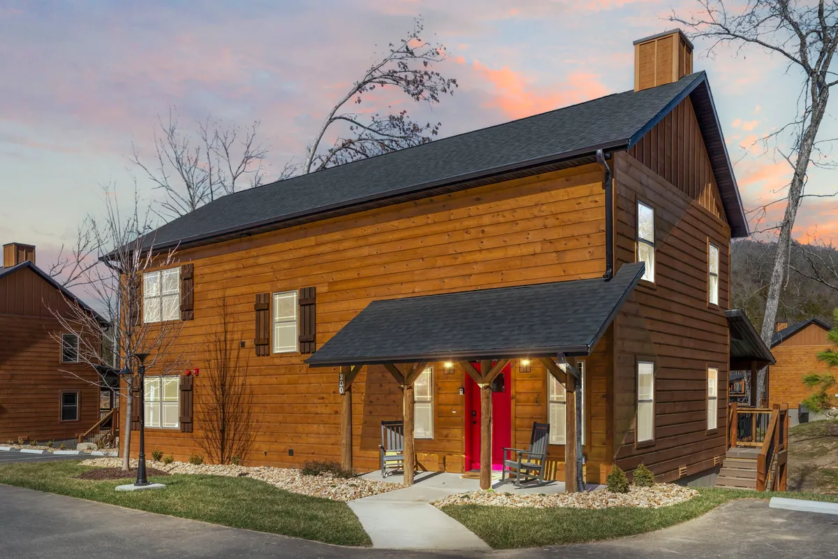 The image showcases the charming wooden exterior of the cabin, featuring a striking red door and a welcoming front porch adorned with classic rocking chairs. The scene is bathed in the warm glow of sunset, inviting guests to relax and enjoy serene views while gathering with loved ones.