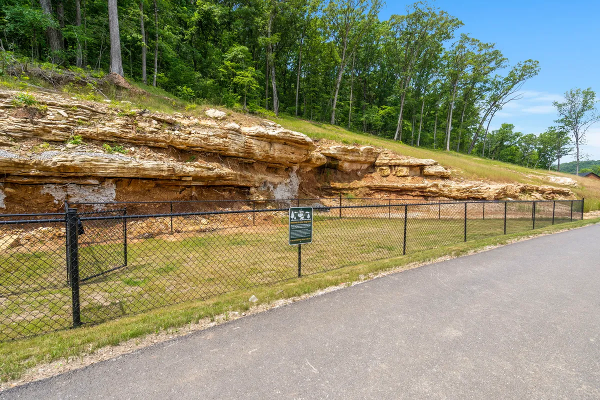 A picturesque hiking trail featuring a unique rock formation, an information sign, and a fence, set within a calm, wooded area. The path is inviting and provides an educational and scenic option for nature walks.