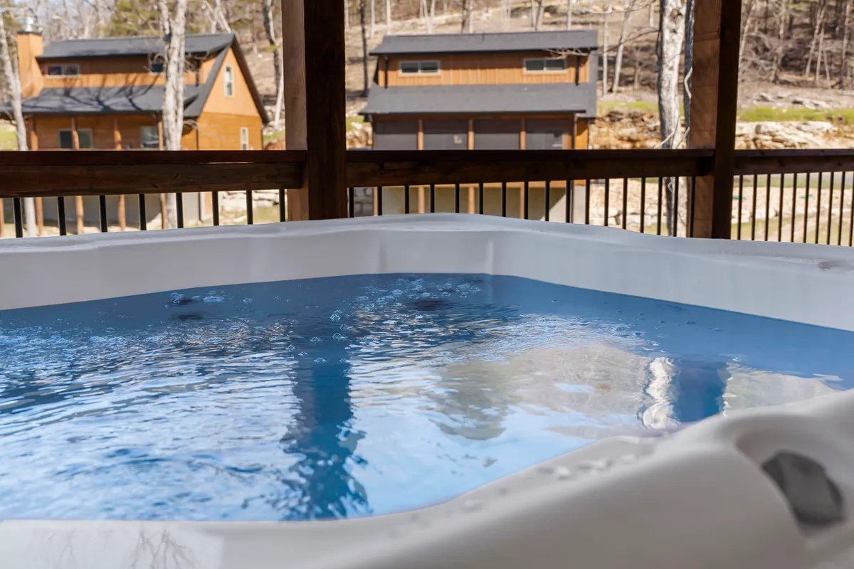 A tranquil outdoor setting highlights a hot tub nestled amid vibrant greenery, with nearby cabins peeking through the trees. The scene invites guests to enjoy both the bubbling hot tub and the natural surroundings, evoking a sense of peaceful seclusion.