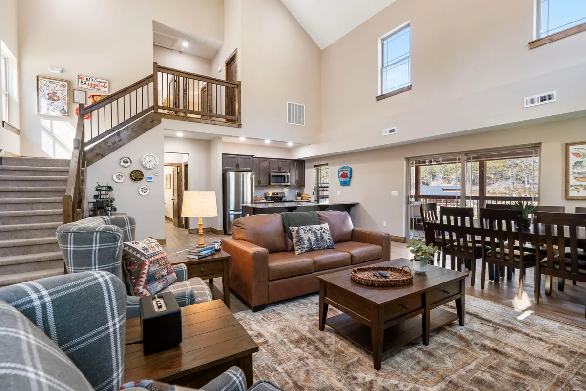 A welcoming open-concept living space featuring a sofa, dining table, staircase, kitchen area, and decorative wall art. The layout seamlessly connects the living, dining, and kitchen areas, emphasizing togetherness and fluid movement between spaces. The warm lighting and inviting decor create a cozy atmosphere for socializing and relaxation.